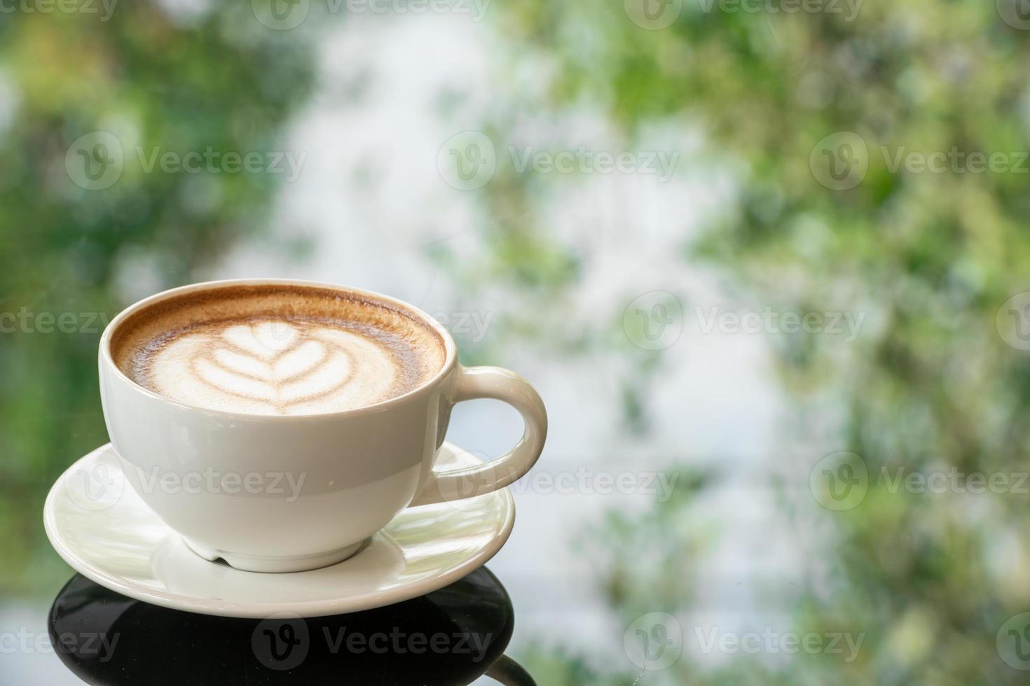 Latte art coffee cup on glass table. Blurred leaf reflection background. photo