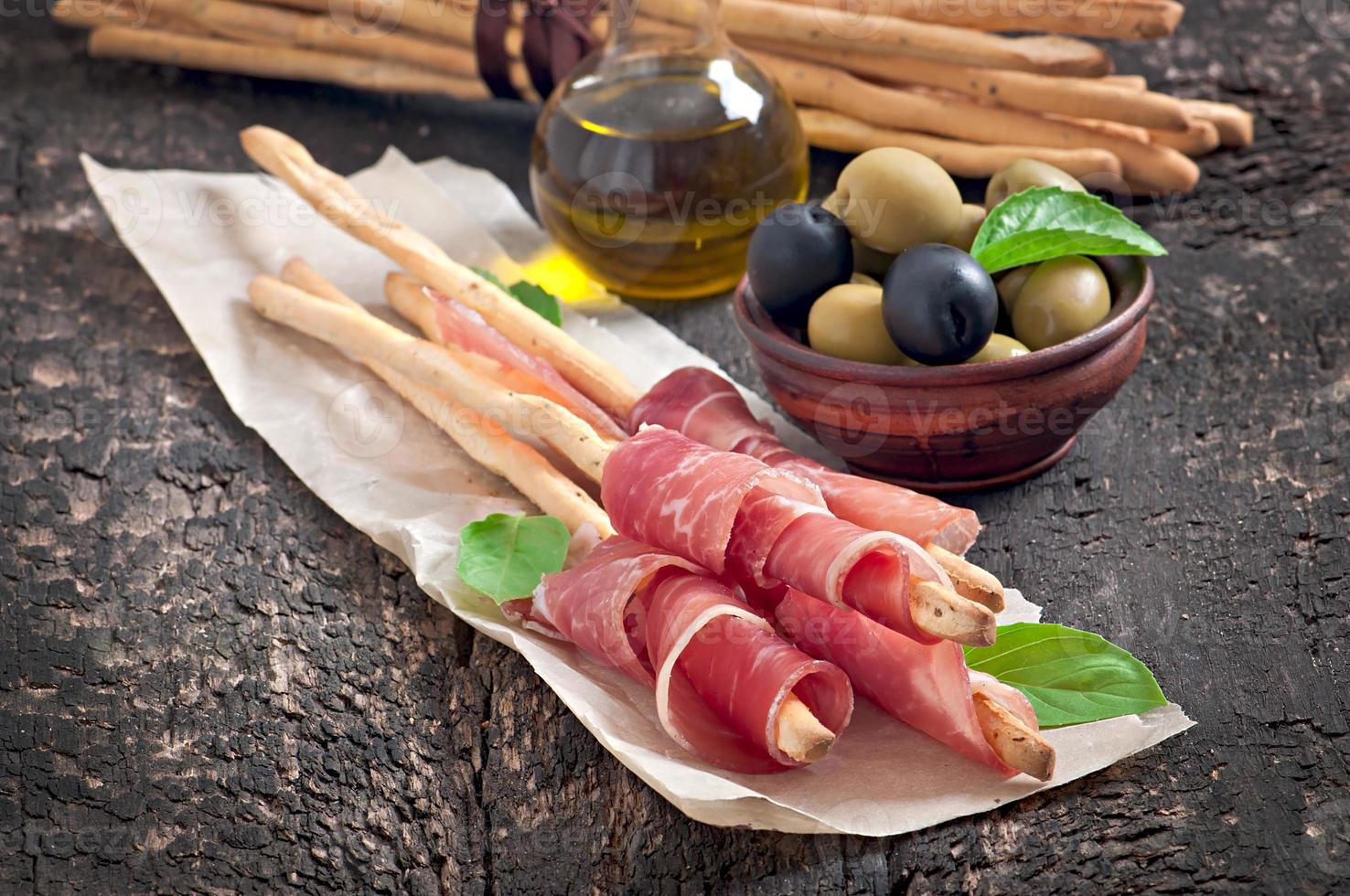 Grissini bread sticks with ham, olives, basil on old wooden background photo