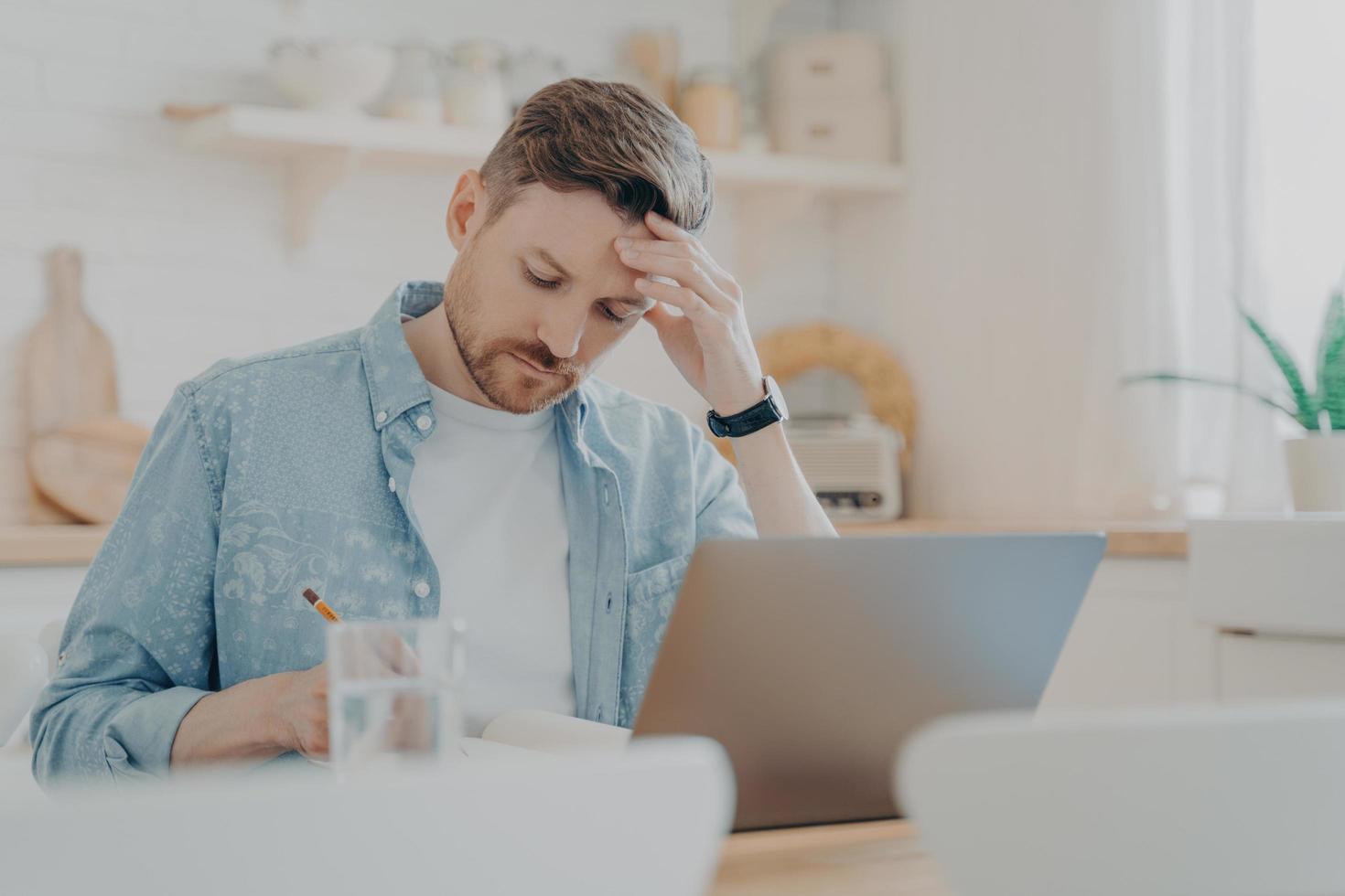 Focused man writing notes while sitting at kitchen table with laptop photo