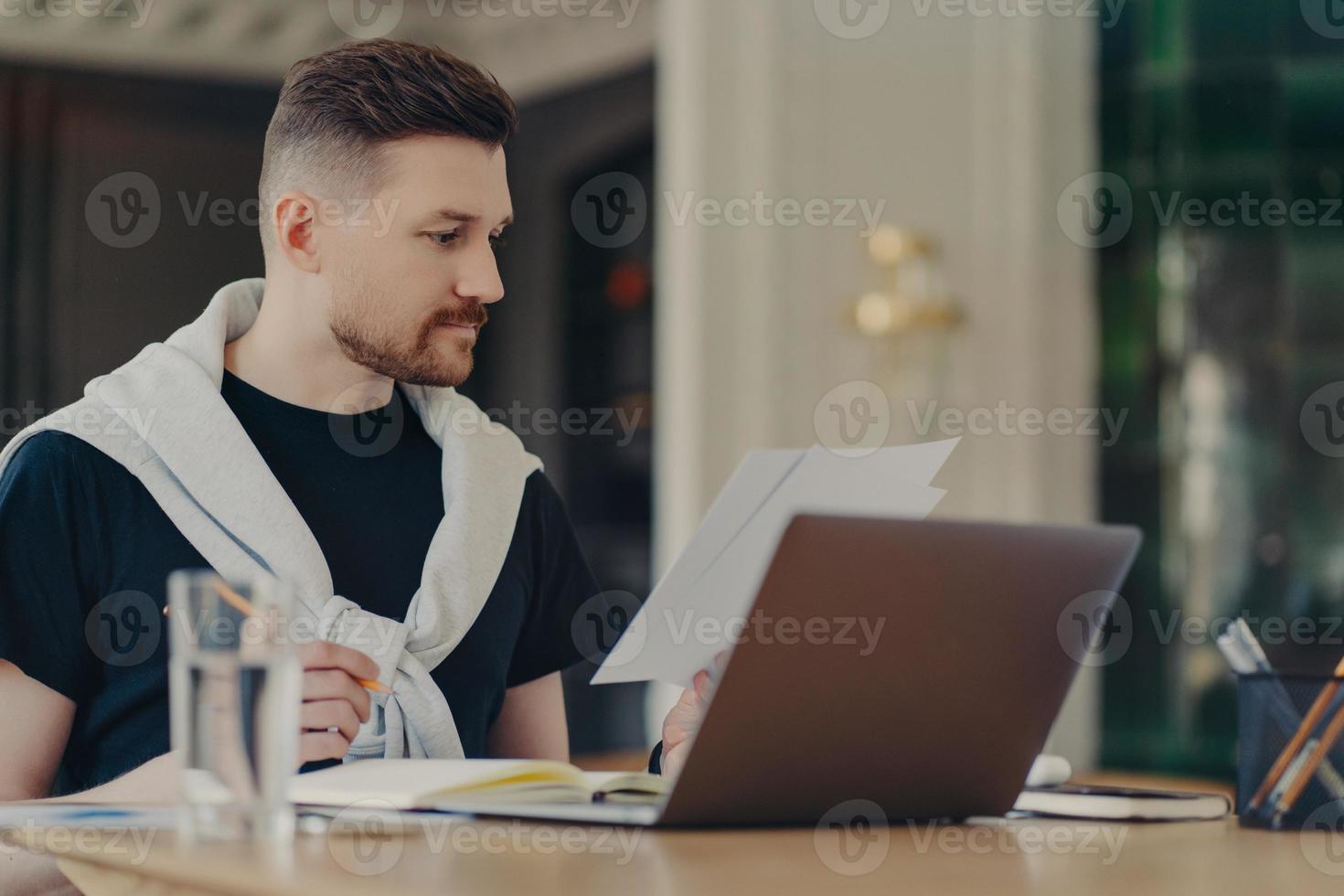 Smart handsome man checks information on papers works remotely sits at home office uses modern laptop computer writes down in notebook dressed in casual clothes. Freelance work and technology photo