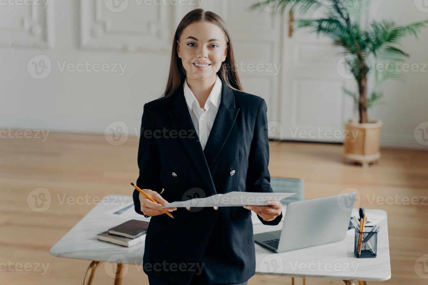 joven mujer de negocios sonriente mirando a la cámara y sosteniendo un documento frente al escritorio de la oficina foto