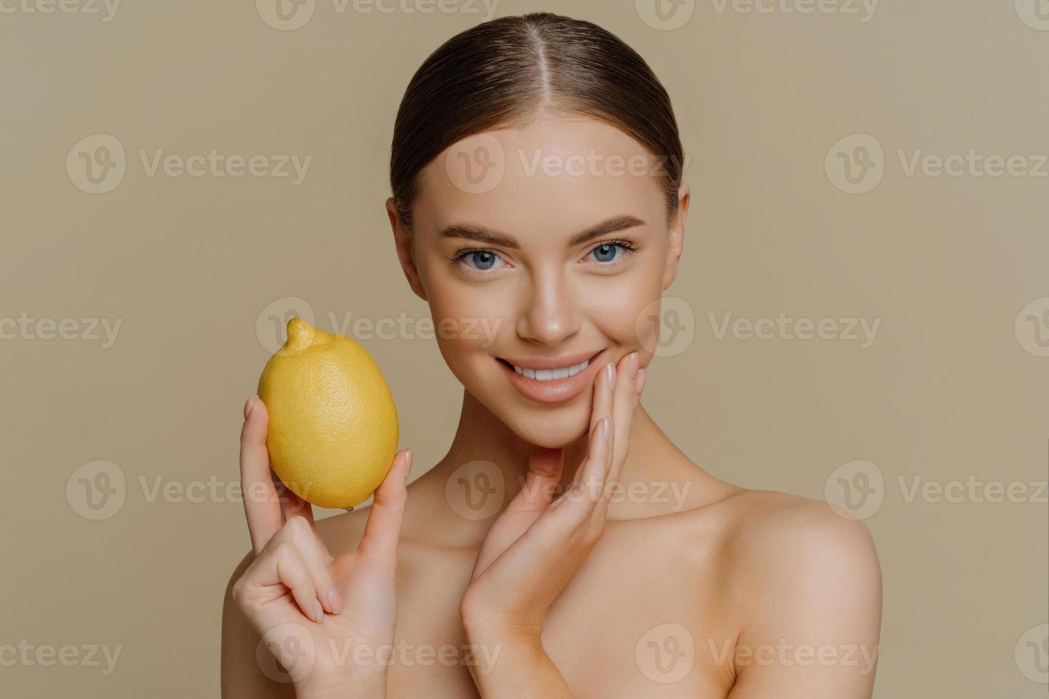 la foto horizontal de una mujer europea bastante saludable usa limón para hacer una mascarilla facial natural que cuida su cuerpo y la piel permanece en topless en el interior aislada sobre un fondo marrón. concepto de belleza.