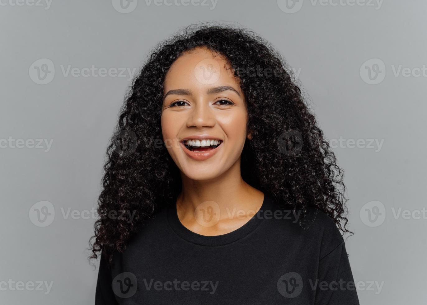 Close up shot of happy dark skinned Afro American woman laughs positively, being in good mood, dressed in black casual clothes, isolated on grey background. Human emotions and feeligs concept. photo