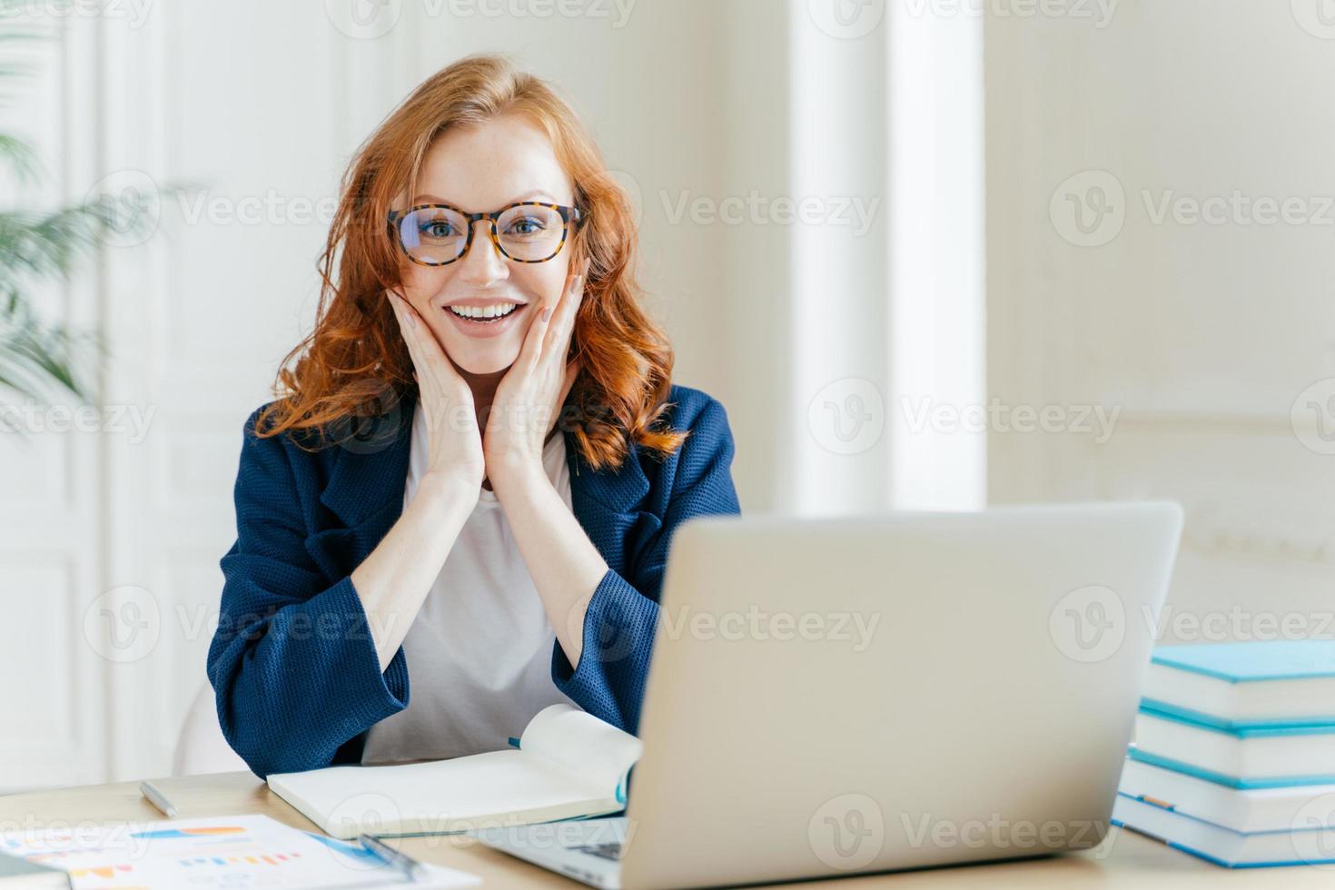 Positive redhead executive manager searches information on internet website, touches both cheeks, writes notes in notepad, uses modern laptop computer, dressed in formal clothes, checks organiser. photo
