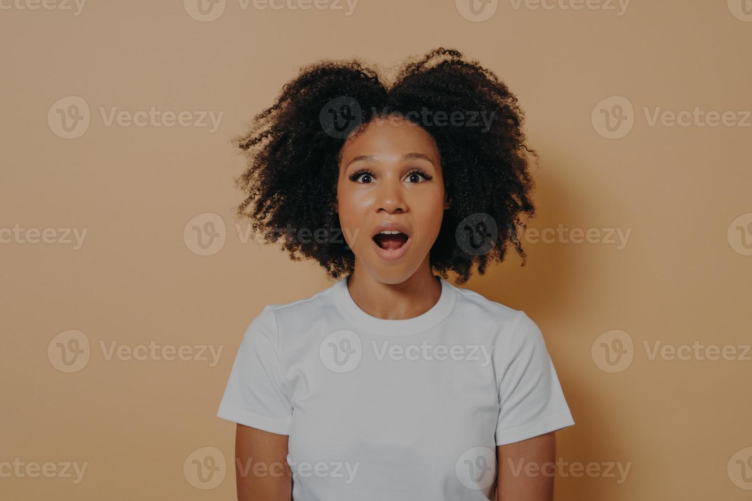 Amazed stunned dark skinned female looking at camera with opened mouth isolated over beige wall photo