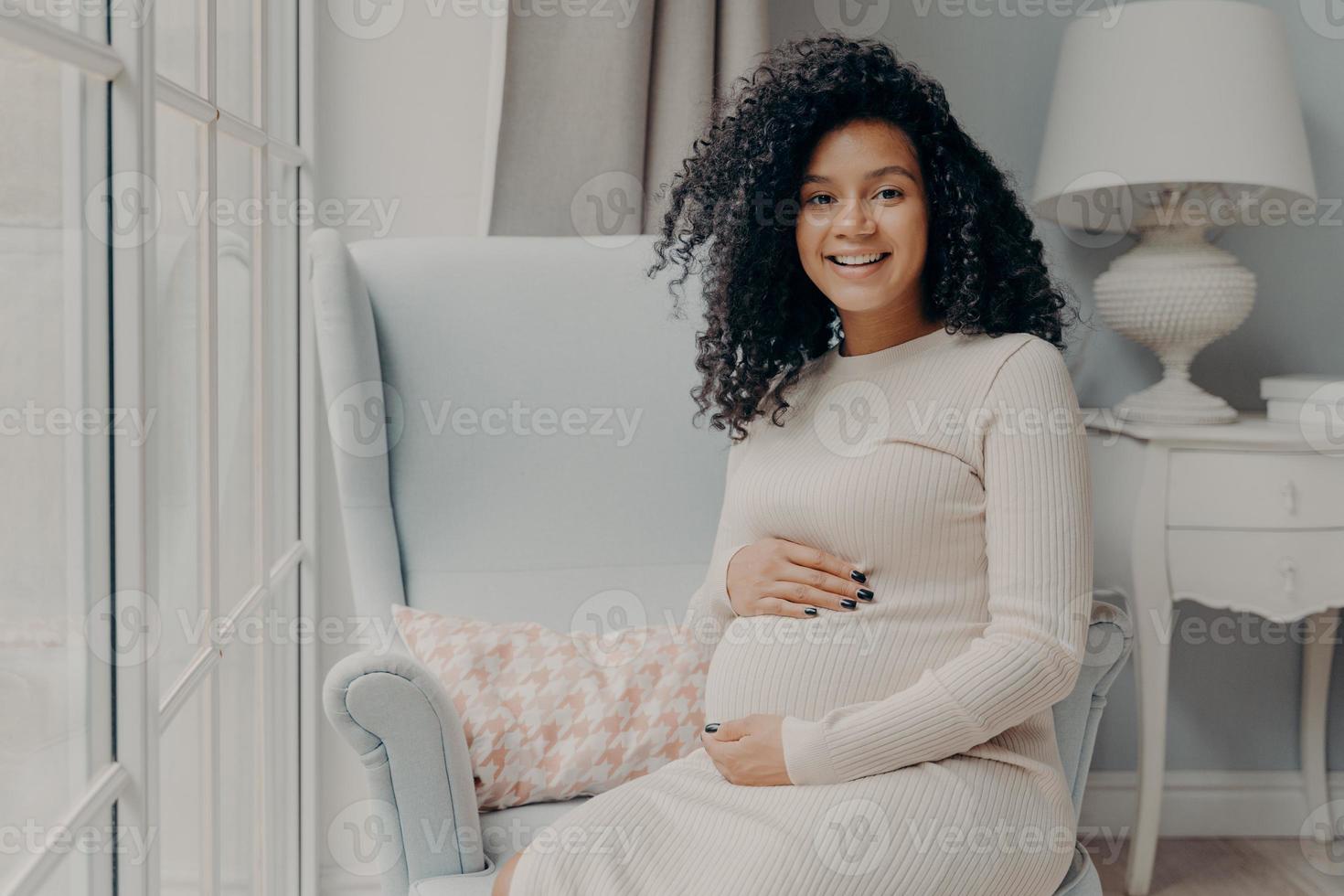 afroamericana adorable mujer embarazada sonriente con vestido beige sentada en un sillón blanco foto