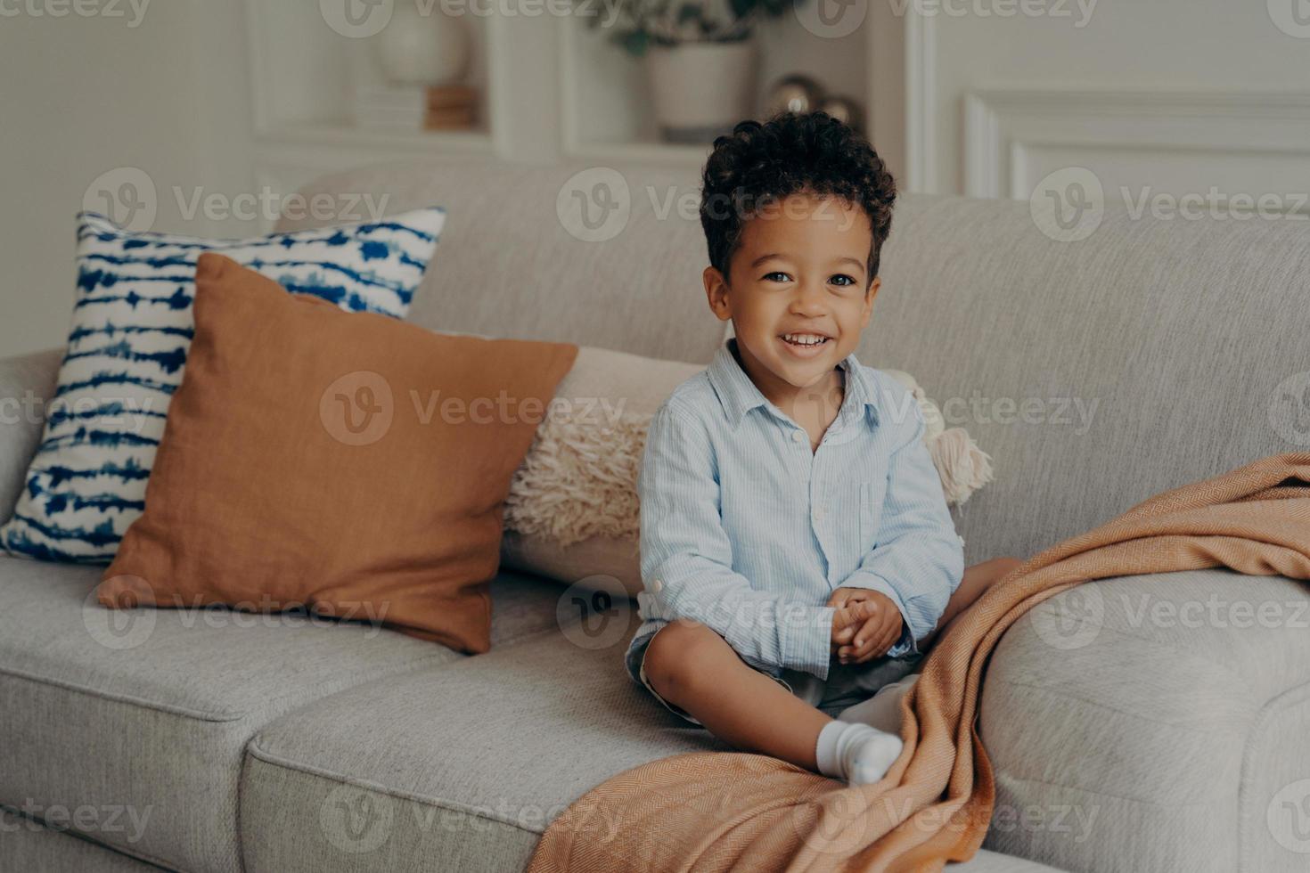 Sweet afro kid boy in casual clothes sitting on comfortable sofa and playing indoors photo