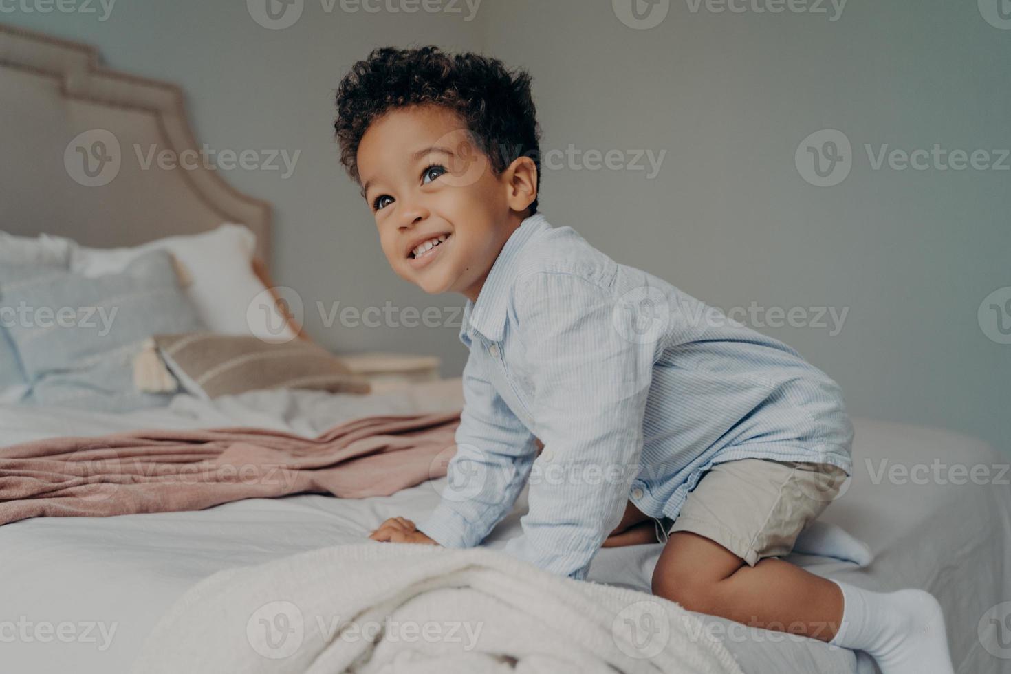 Smiling Afro American child trying to climb on big bed at home, having fun indoors photo