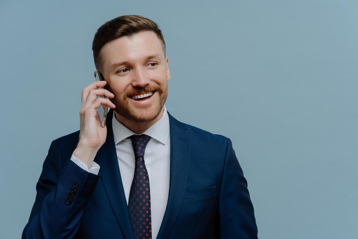 Glad male employer has telephone conversation with colleague looks away smiles gladfully wears formal suit and tie connected to roaming while having business trip poses against blue background photo