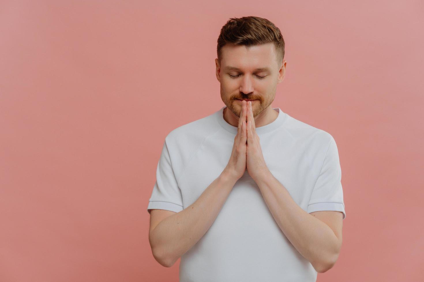 Young bearded man with closed eyes keeping hands in praying gesture photo