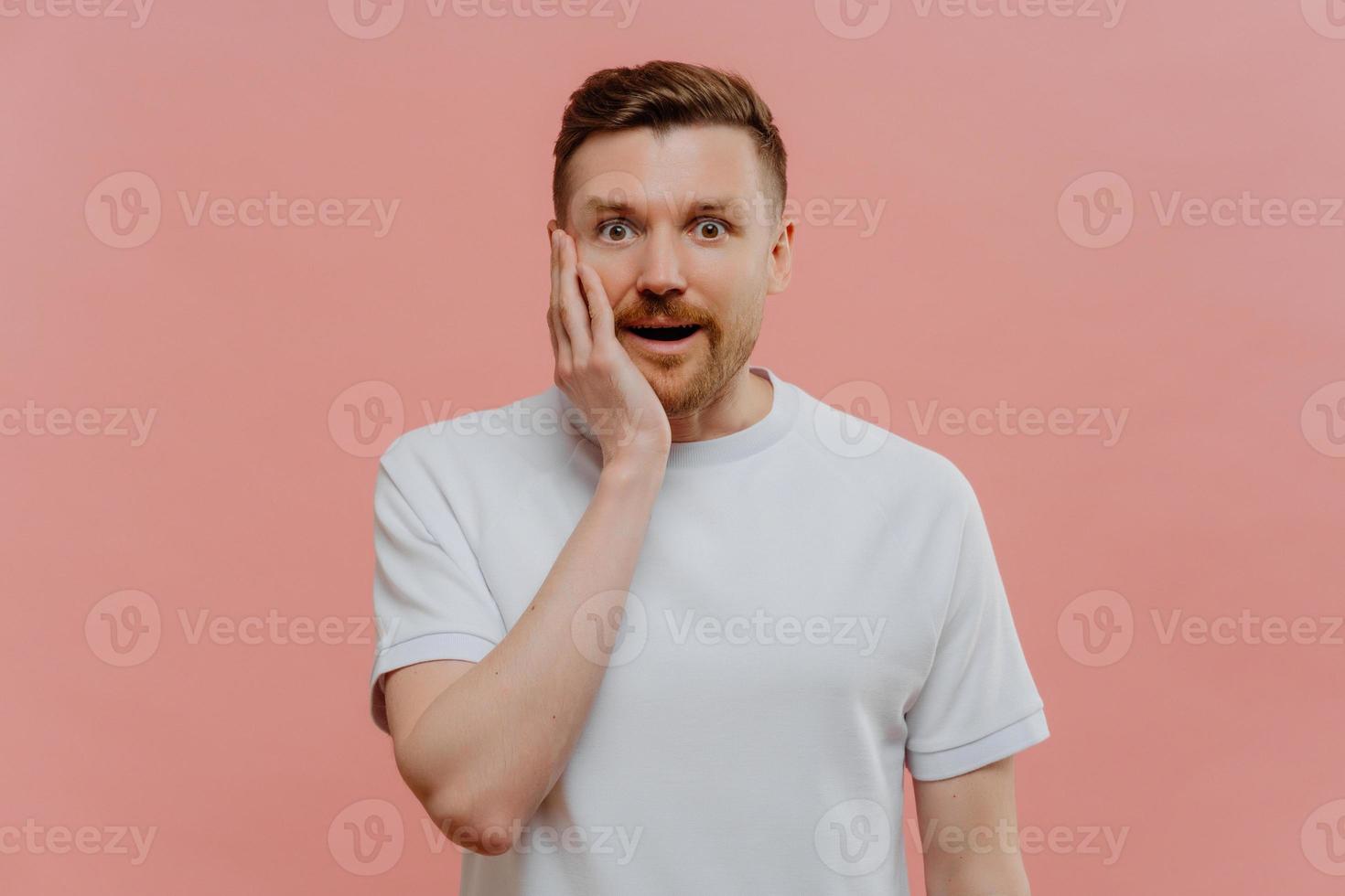 Amazed bearded man keeps hand on cheek stands speechless cannot believe his eyes dressed in casual white t shirt isolated over pink background. Startled unshaven male model finds out shocking news photo
