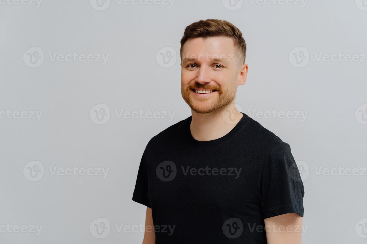 Portrait of attractive European man smiles carefree being in good mood stands satisfied dressed in casual black t shirt poses against blue background copy space for your advertising content. photo