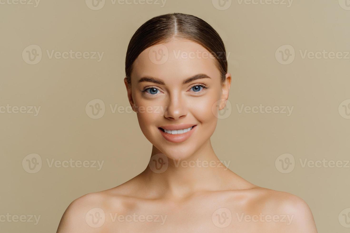 Beauty portrait of charming European woman with beautiful face healthy complexion well cared body smiles gently stands with bare shoulders indoor against beige background has clean fresh skin photo