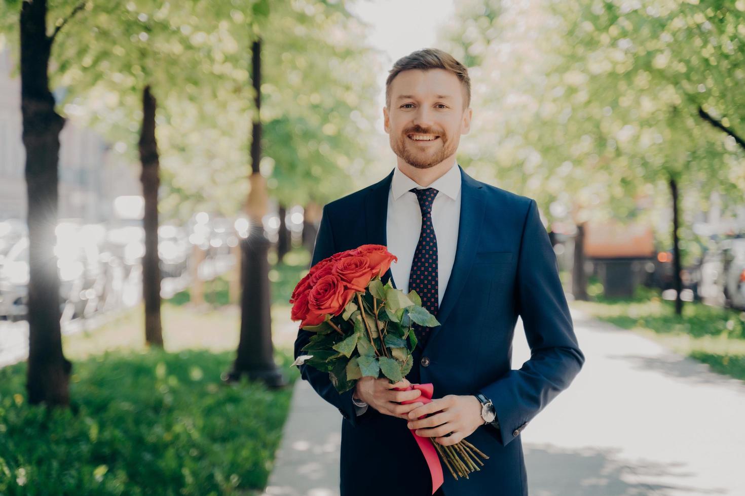 Happy young man in elegant suit with bouquet of flowers walking in park photo