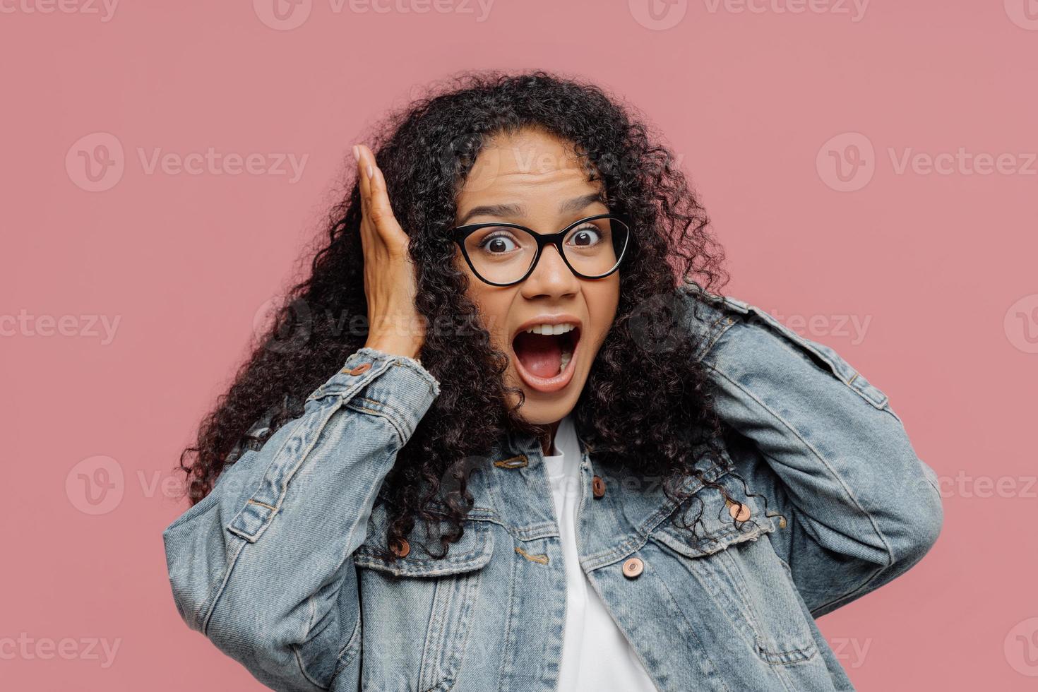 Photo of surprised Afro American woman covers ears, yells loudly, ignores loud sound, keeps mouth widely opened, wears spectacles and denim jacket, stands against rosy wall. Omg, how noisy there