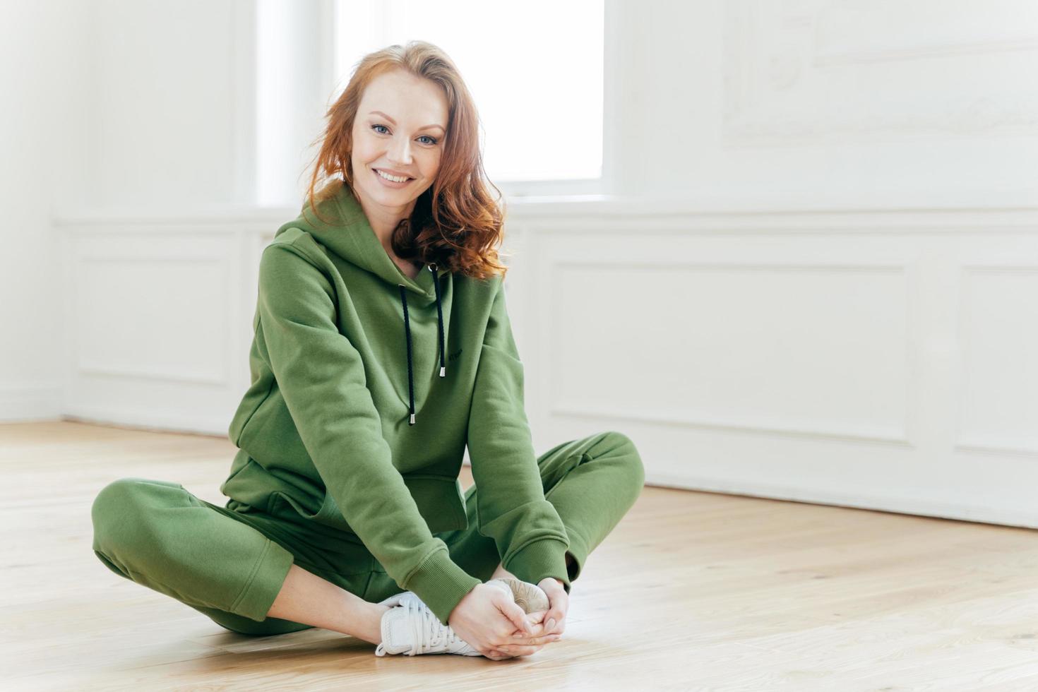 Indoor shot of beautiful redhead European woman has rest after cardio training, keeps legs crossed, dressed in green tracksuit, sits on floor alone. Healthy lifestyle, youth and fitness concept. photo