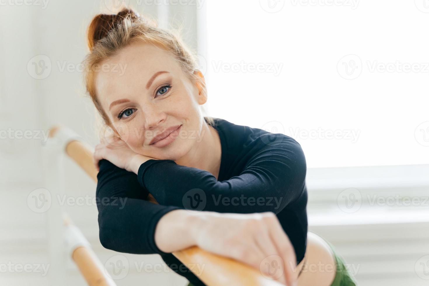 Close up portrait of ginger attractive female dancer with combed hair, makeup, wears black top, leans on barre, has gentle smile, looks straightly at camera. People, choreography, leisure concept photo