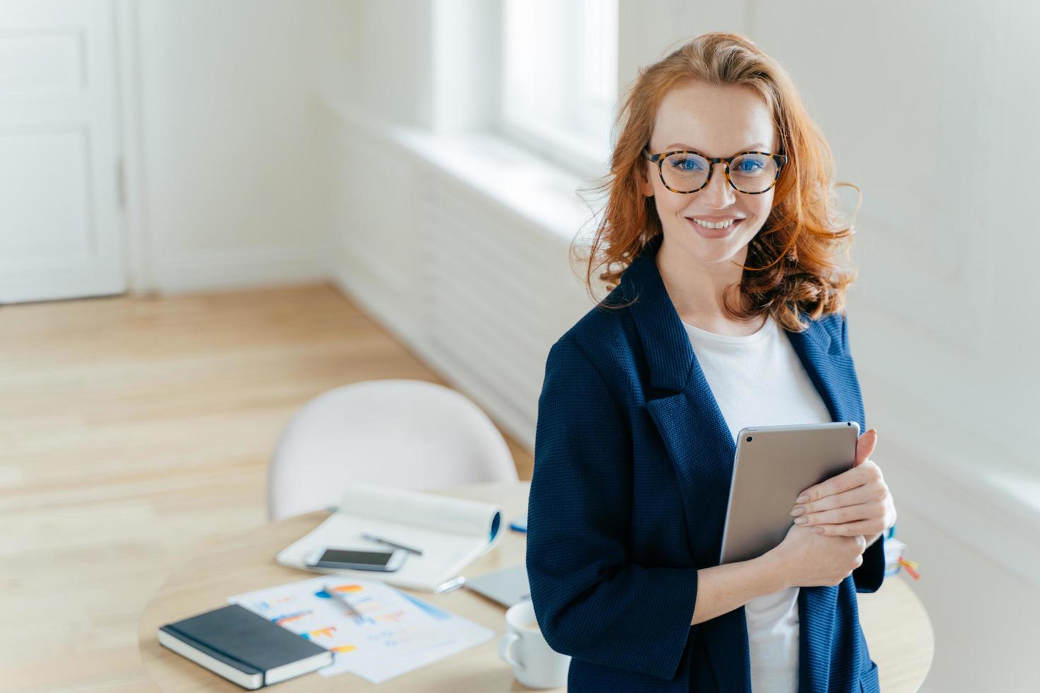 Prosperous female leader of working team holds digital tablet device, develops business ideas, has toothy smile, red hair, wears elegant outfit, stands in own cabinet, involved in working process photo