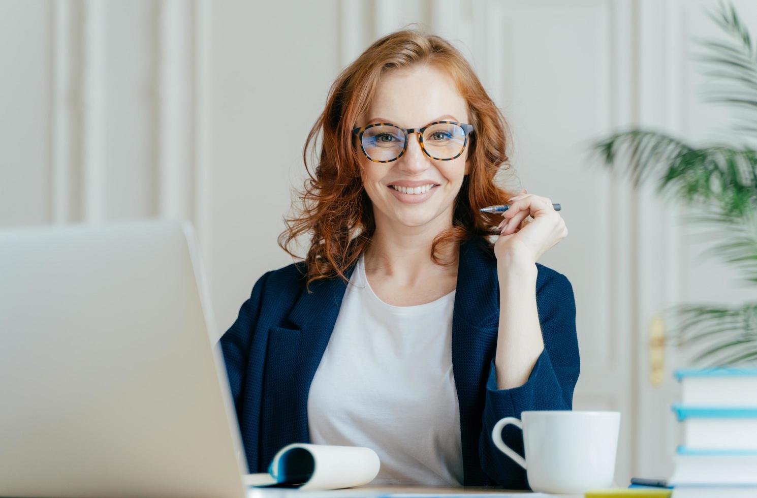 Glad redhead curly woman works freelance, uses laptop computer, writes down notes in notepad, drinks coffee, wears optical glasses, being in good mood, develops business company. Work concept photo