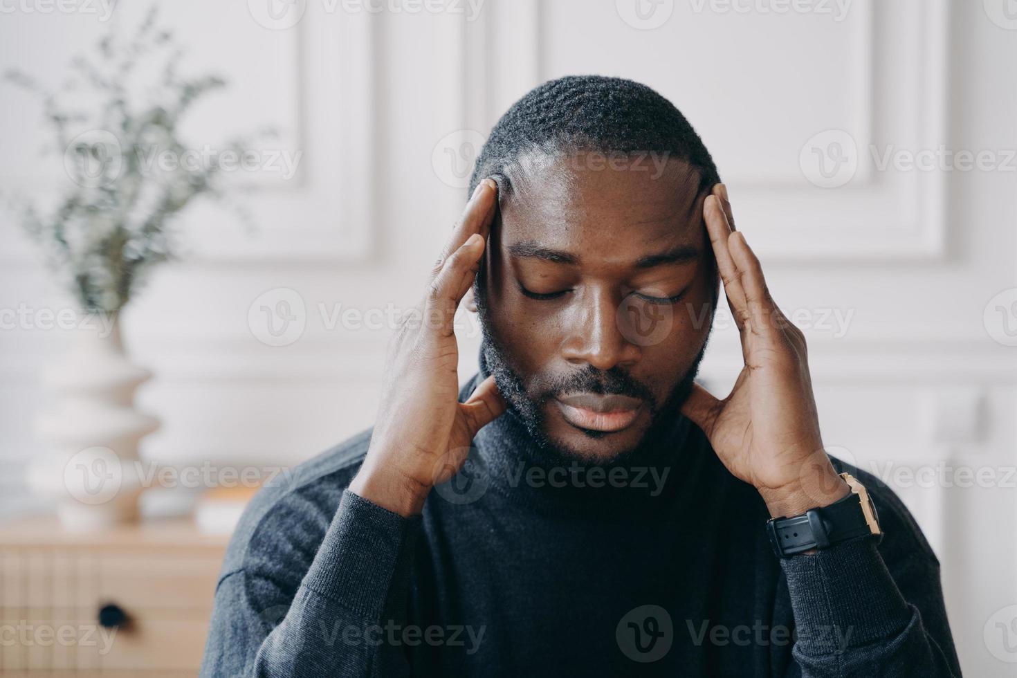 agotado joven empresario afroamericano sentado con los ojos cerrados y con las manos en la cabeza foto