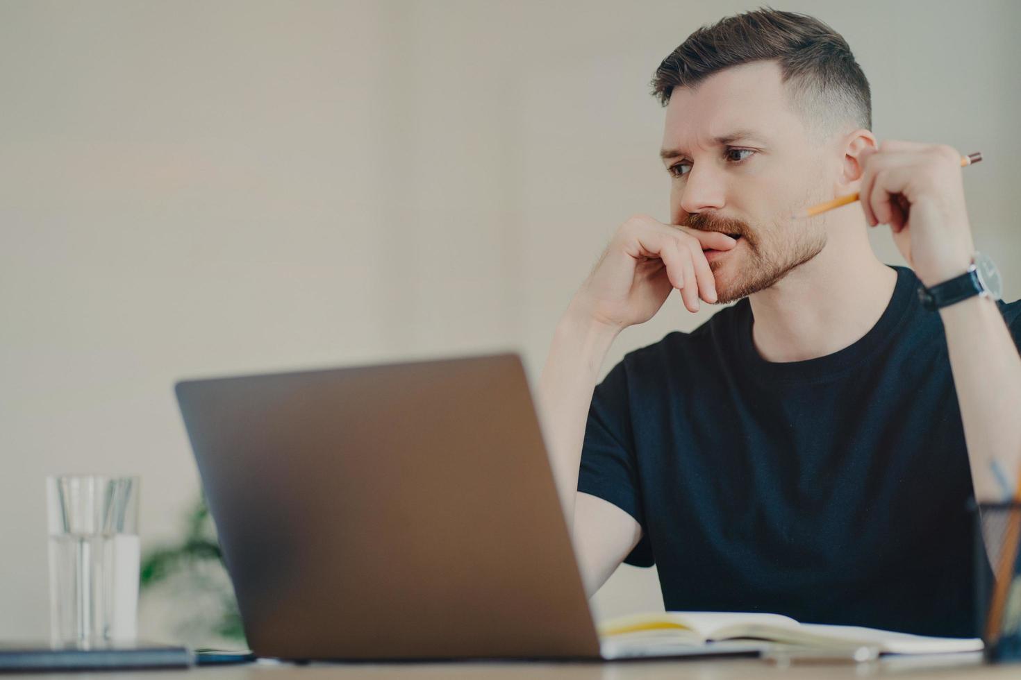 Serious bearded man student enjoys studying process has thoughtful expression works freelance dressed in casual black t shirt writes down notes uses modern laptop computer poses in coworking space photo