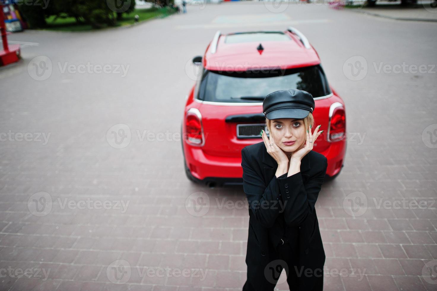 retrato de una hermosa modelo de mujer rubia sexy con gorra y todo negro con maquillaje brillante cerca del coche rojo de la ciudad. foto