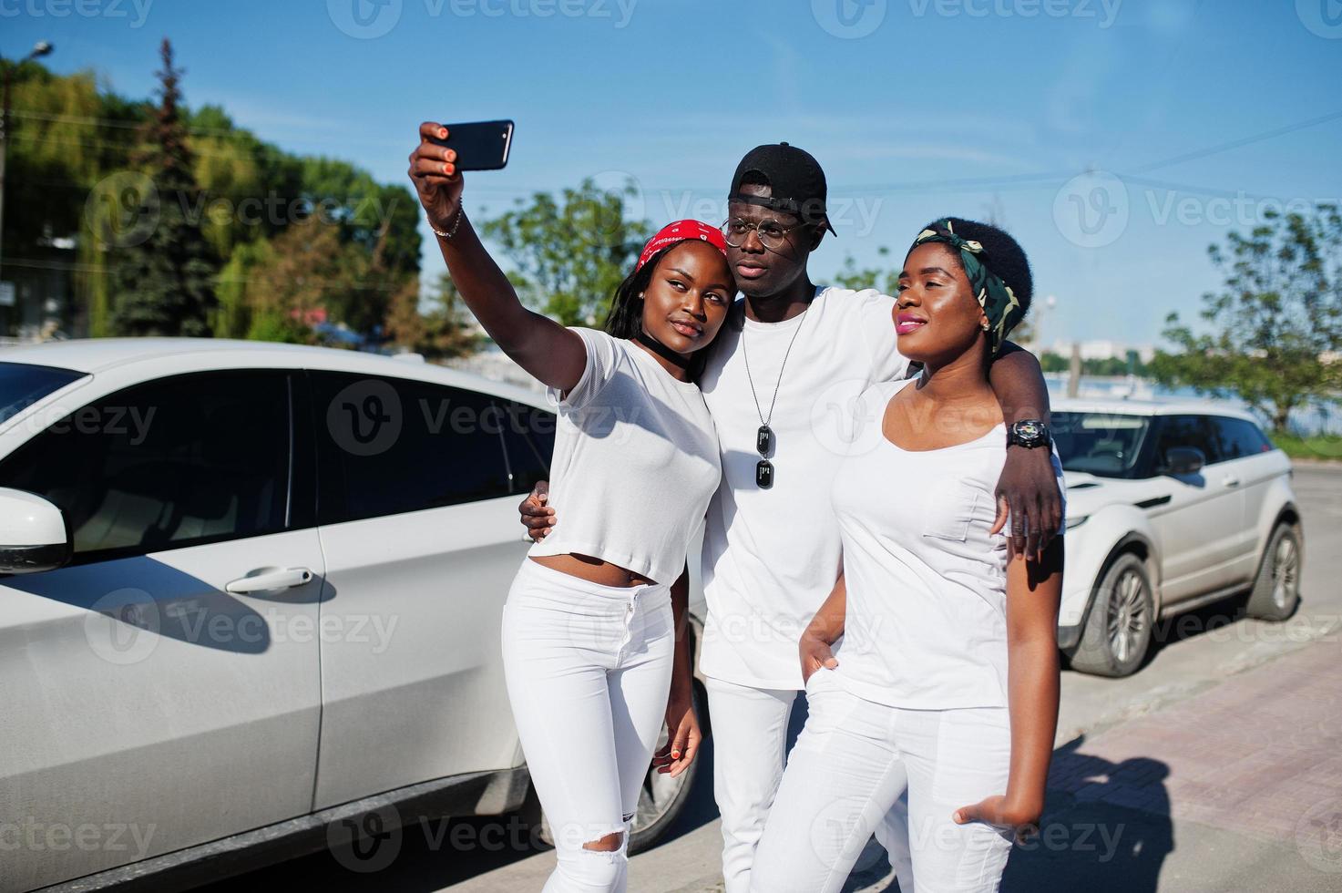 Three stylish african american friends making selfie on phone, wear on white clothes against two luxury cars. Street fashion of young black people. Black man with two african girls. photo