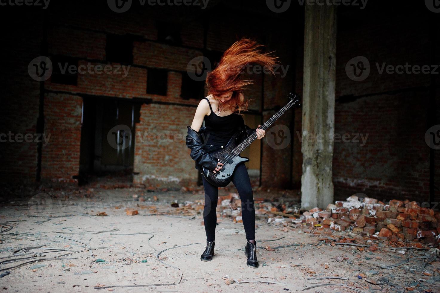 Red haired punk girl wear on black with bass guitar at abadoned place. Portrait of gothic woman musician. photo