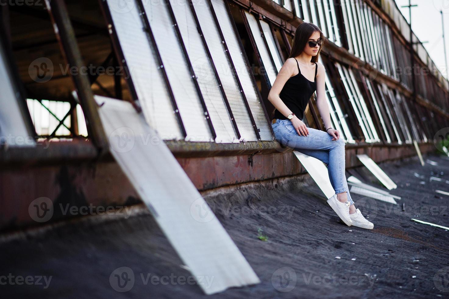 Girl at sunglasses and jeans posed at the roof of abadoned industrial place with windows. photo