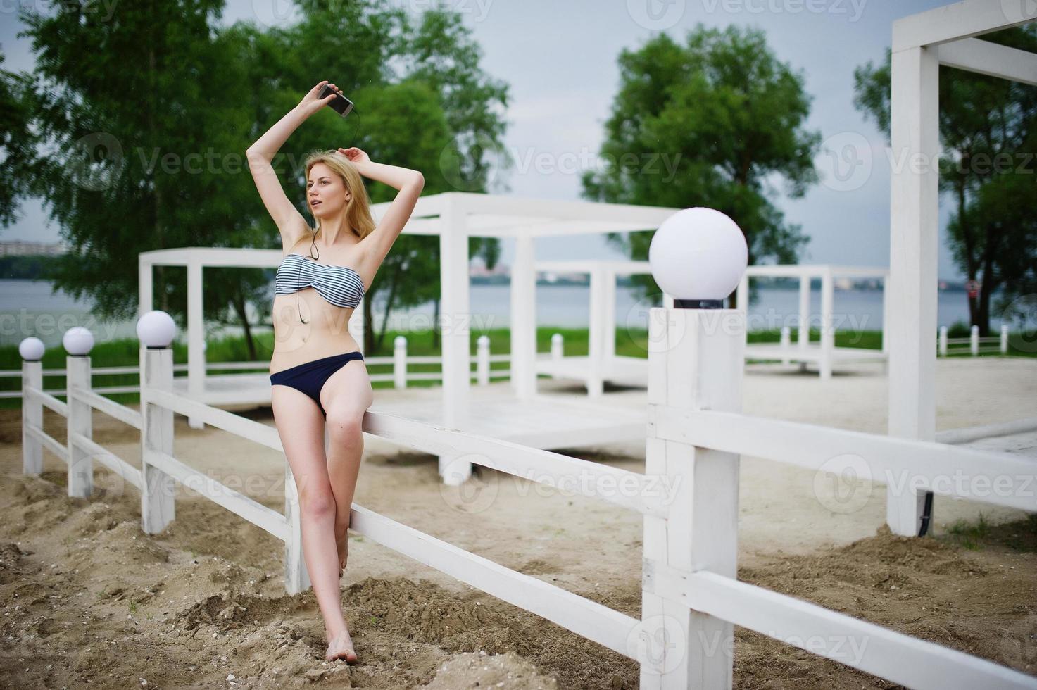 Portrait of a stunning young female model in bikini posing next to the white wooden fence in the park. photo