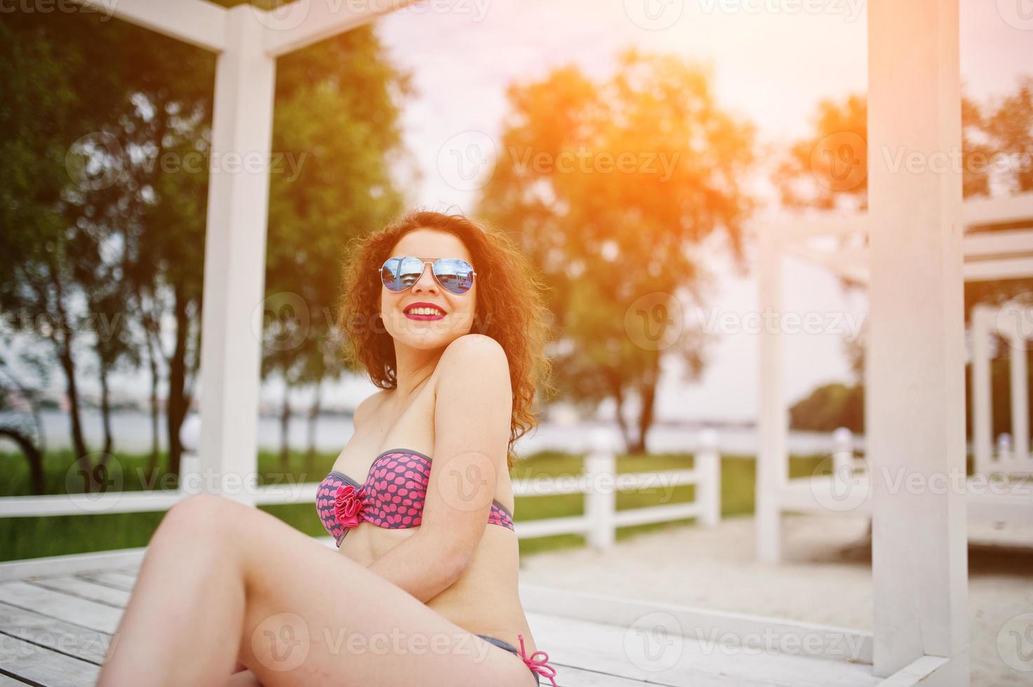 Portrait of an attractive girl posing in bikini with sunglasses on while standing on a white wooden terrace. photo