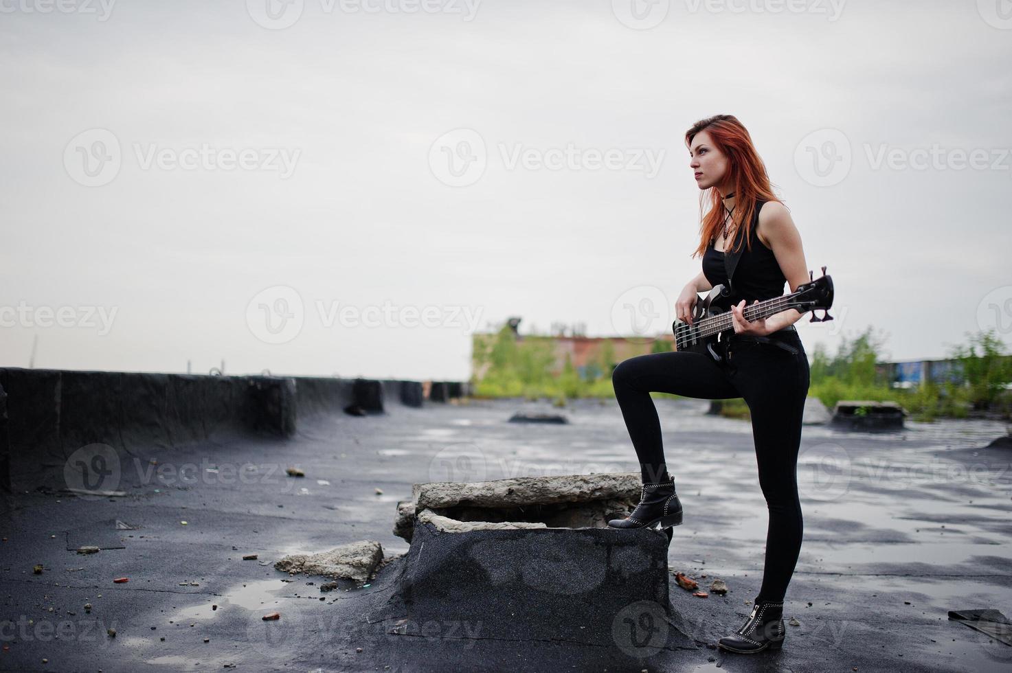 Red haired punk girl wear on black with bass guitar at the roof. photo