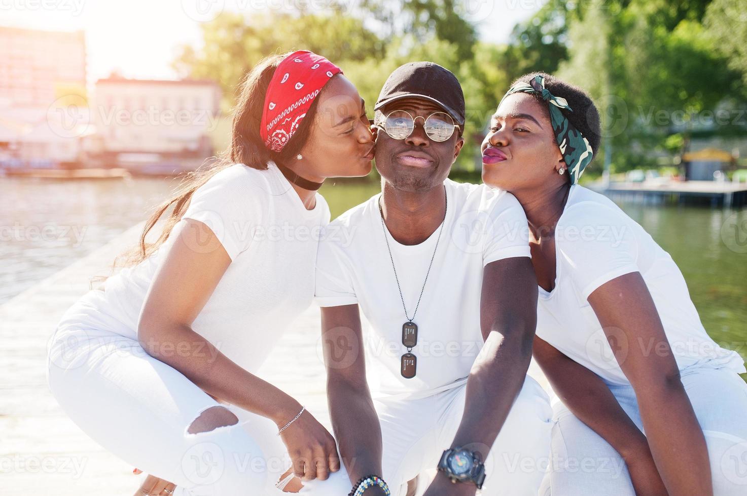 Three stylish african american friends, wear on white clothes at pier on beach. Street fashion of young black people. Black man with two african girls kissed him and show lips. photo