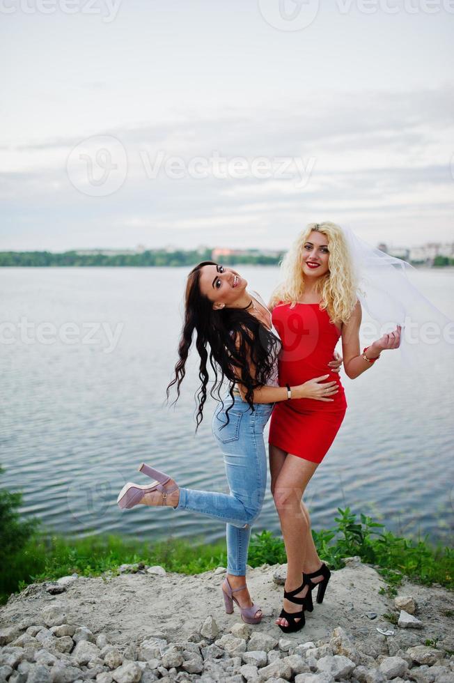 Awesome bride in red dress posing with her bridesmaid on the lakeside. photo