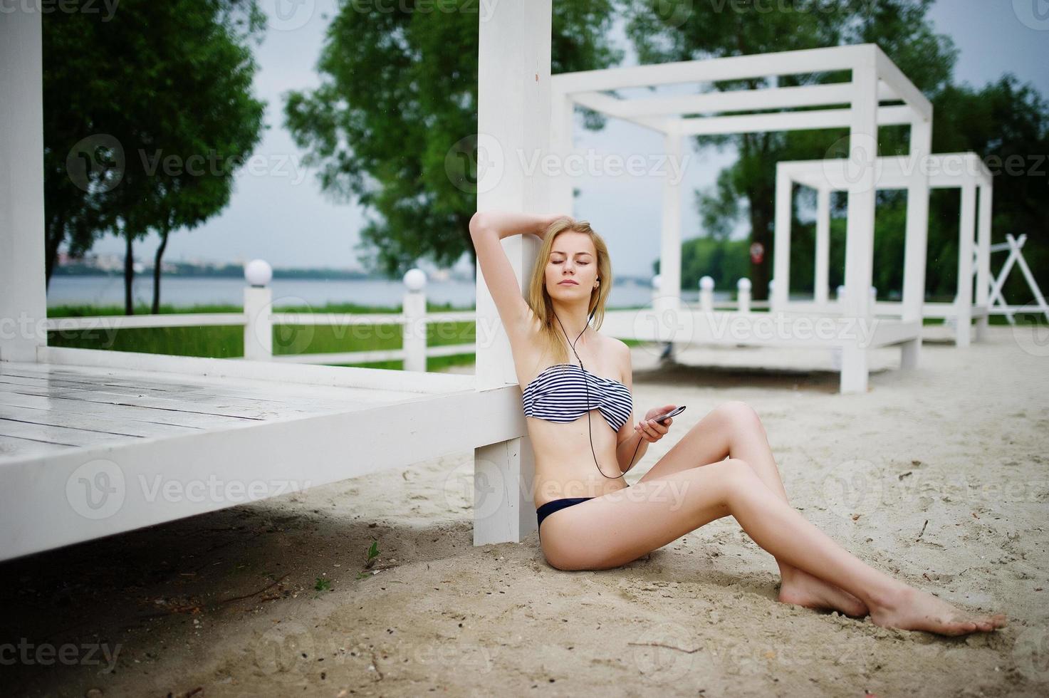 Portrait of a gorgeous young girl in bikini sitting next to the gasebo on sand and posing. photo