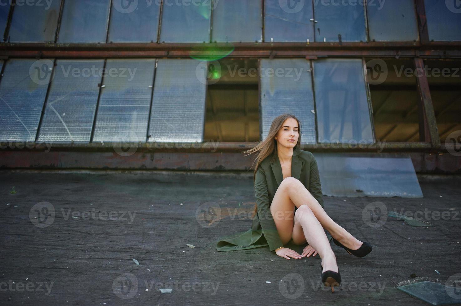 chica modelo sexy con piernas largas en traje de baño de cuerpo de lencería negra vestido combinado y chaqueta posada en el techo de un lugar industrial abandonado con ventanas. foto