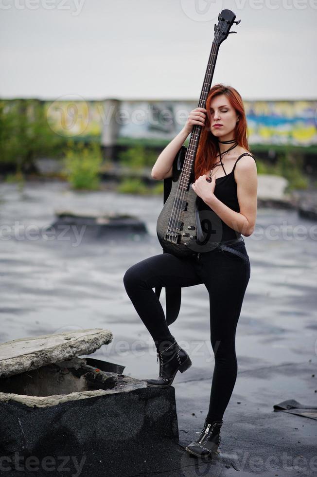 Red haired punk girl wear on black with bass guitar at the roof. photo