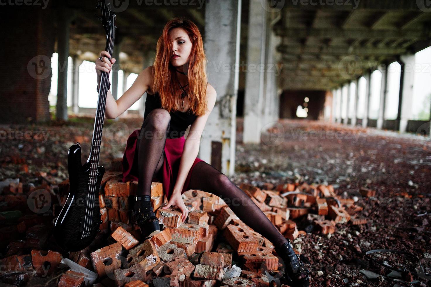 Red haired punk girl wear on black and red skirt, with bass guitar at abadoned place. Portrait of gothic woman musician. photo