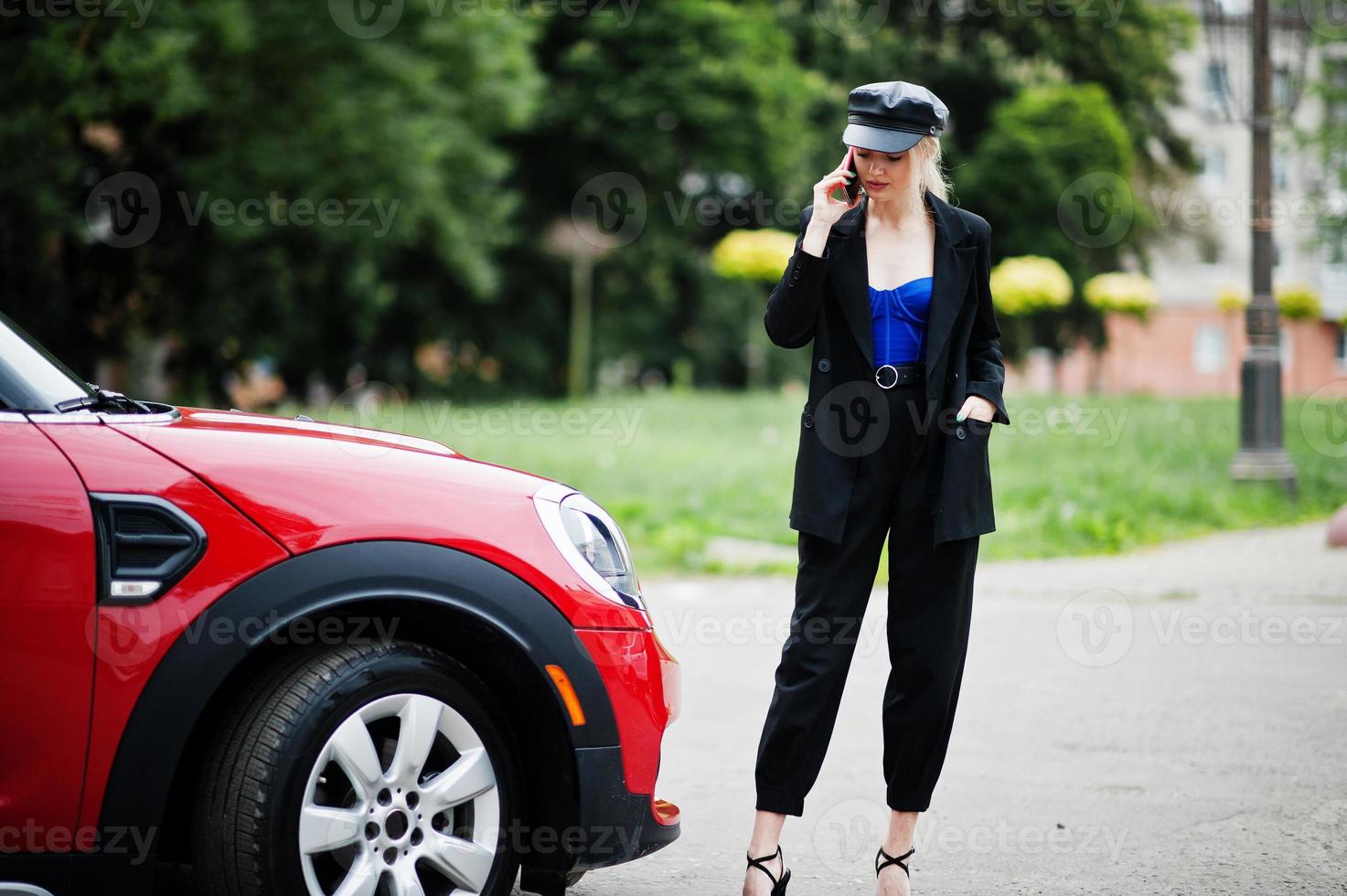 Portrait of beautiful blonde sexy fashion woman model in cap and in all black with bright makeup near red city car, with mobile phone. photo