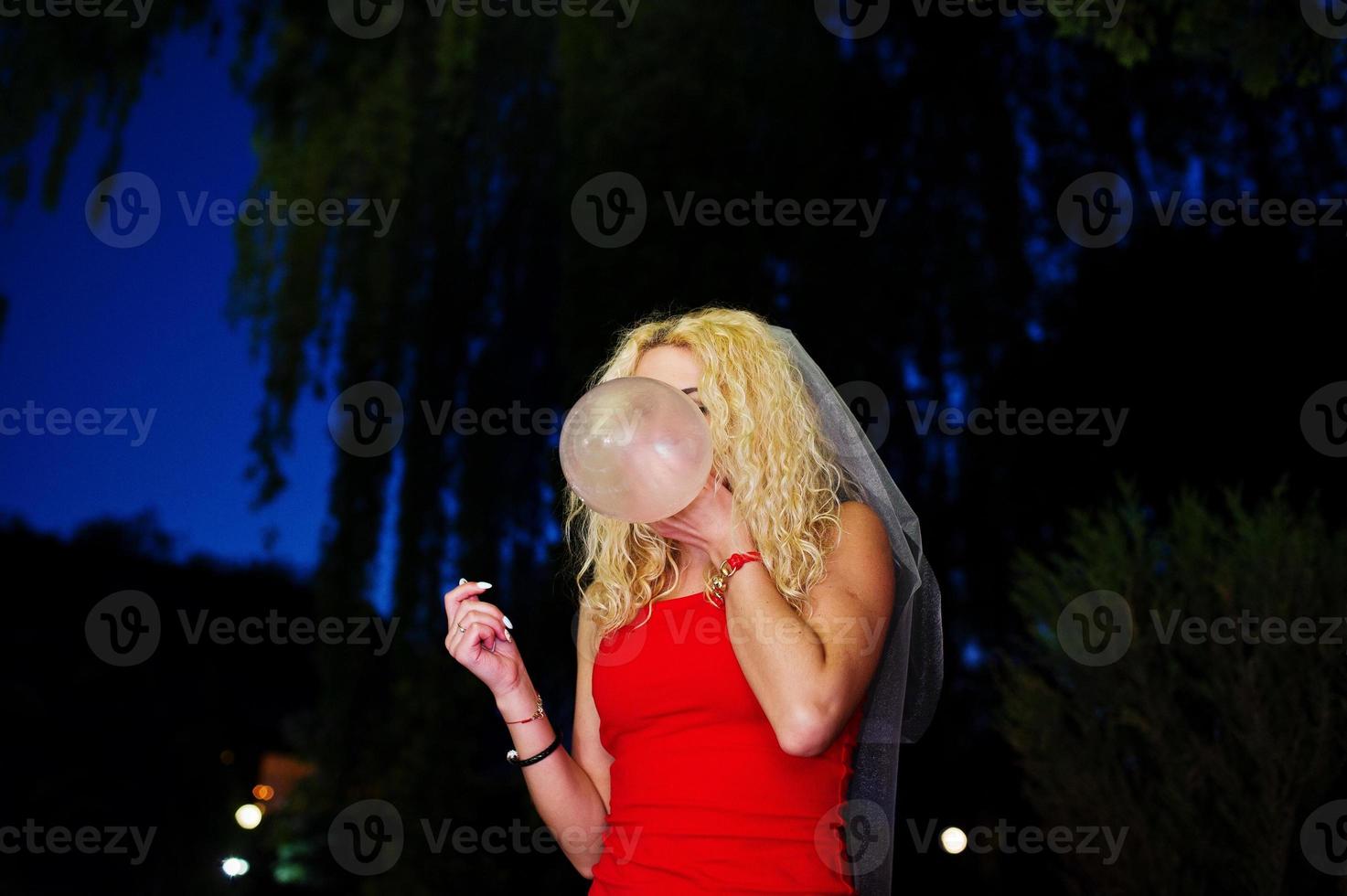 Crazy bride in red dress and veil inflating condom at her bachelorette party. photo