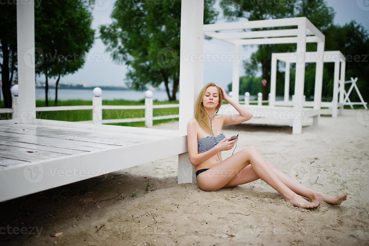 Portrait of a gorgeous young girl in bikini sitting next to the gasebo on sand and posing. photo