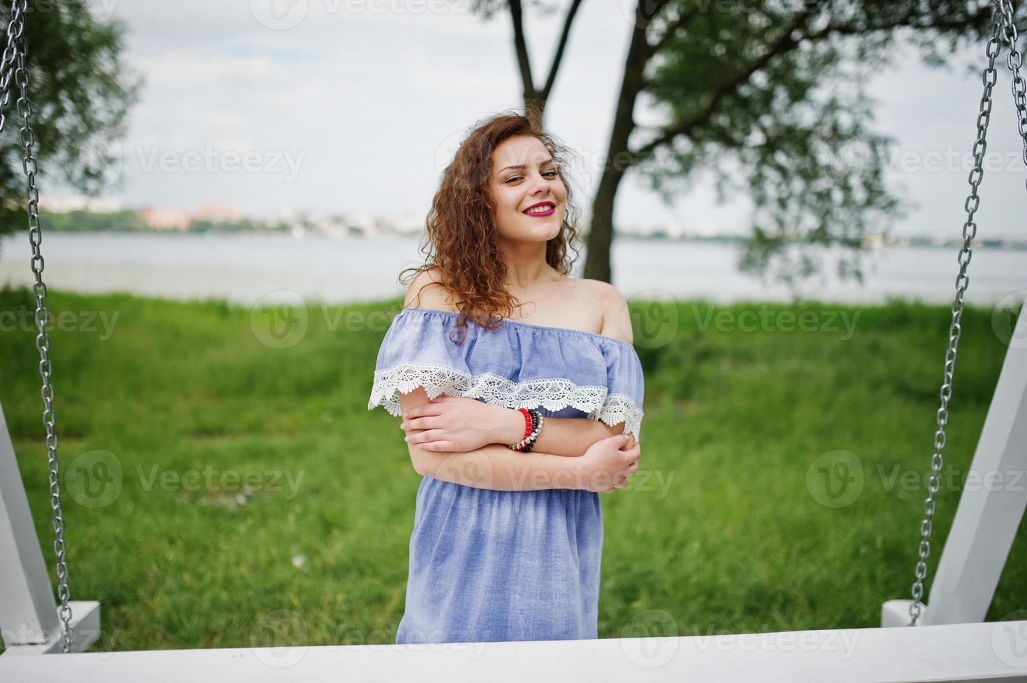 Portrait of a stunning young girl wearing blue marine-styled dress sitting on swings on the lakeside. photo