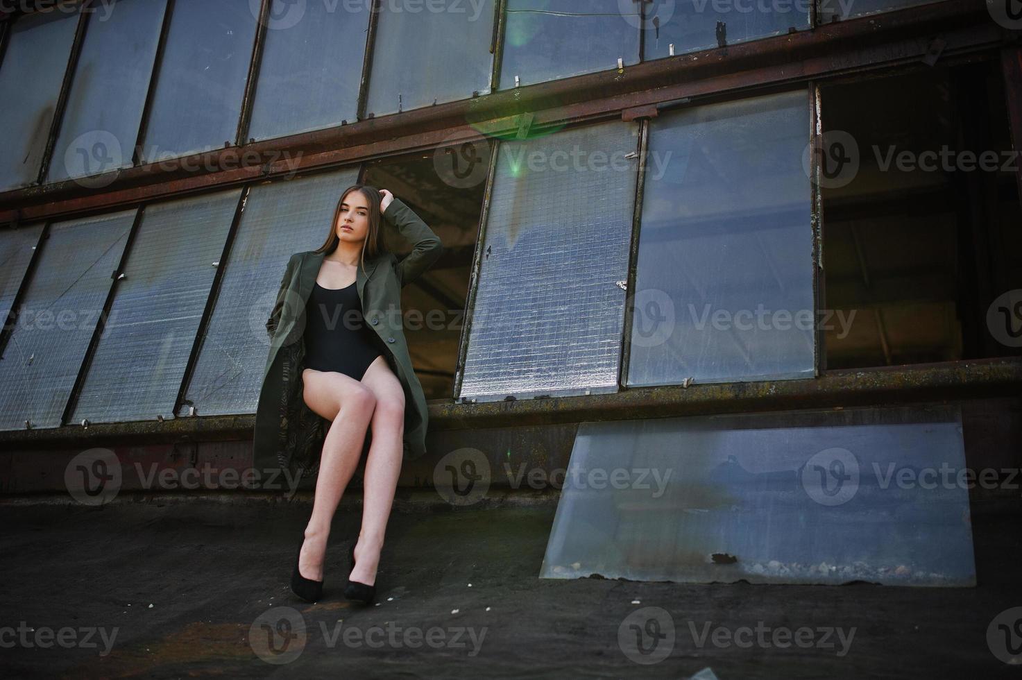 chica modelo sexy con piernas largas en traje de baño de cuerpo de lencería negra vestido combinado y chaqueta posada en el techo de un lugar industrial abandonado con ventanas. foto