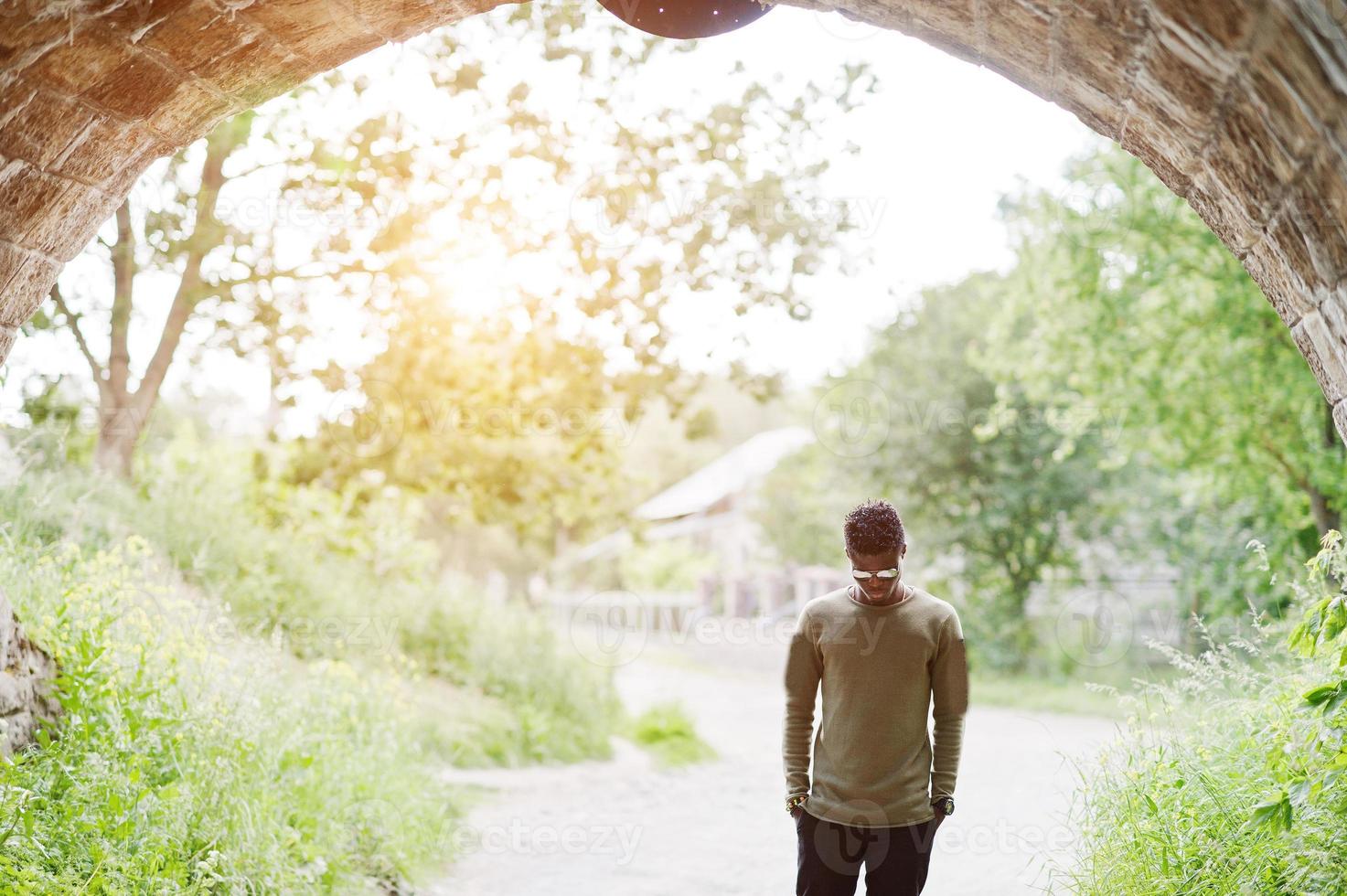 apuesto joven afroamericano con ropa informal y gafas de sol posando y caminando en el túnel. foto