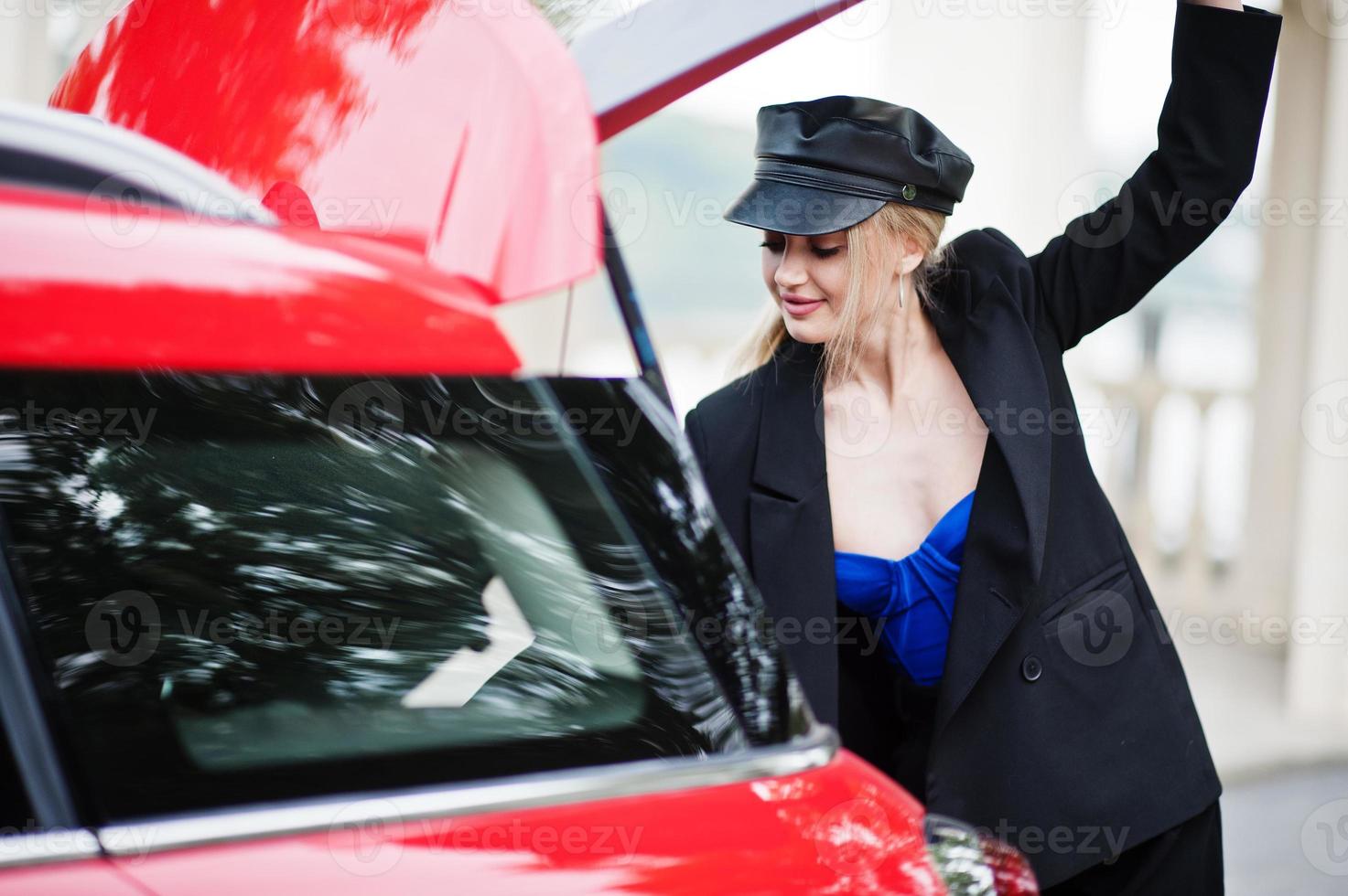 retrato de una hermosa modelo rubia de moda sexy con gorra y todo negro con maquillaje brillante cerca del auto rojo de la ciudad puso su bolso en el baúl. foto