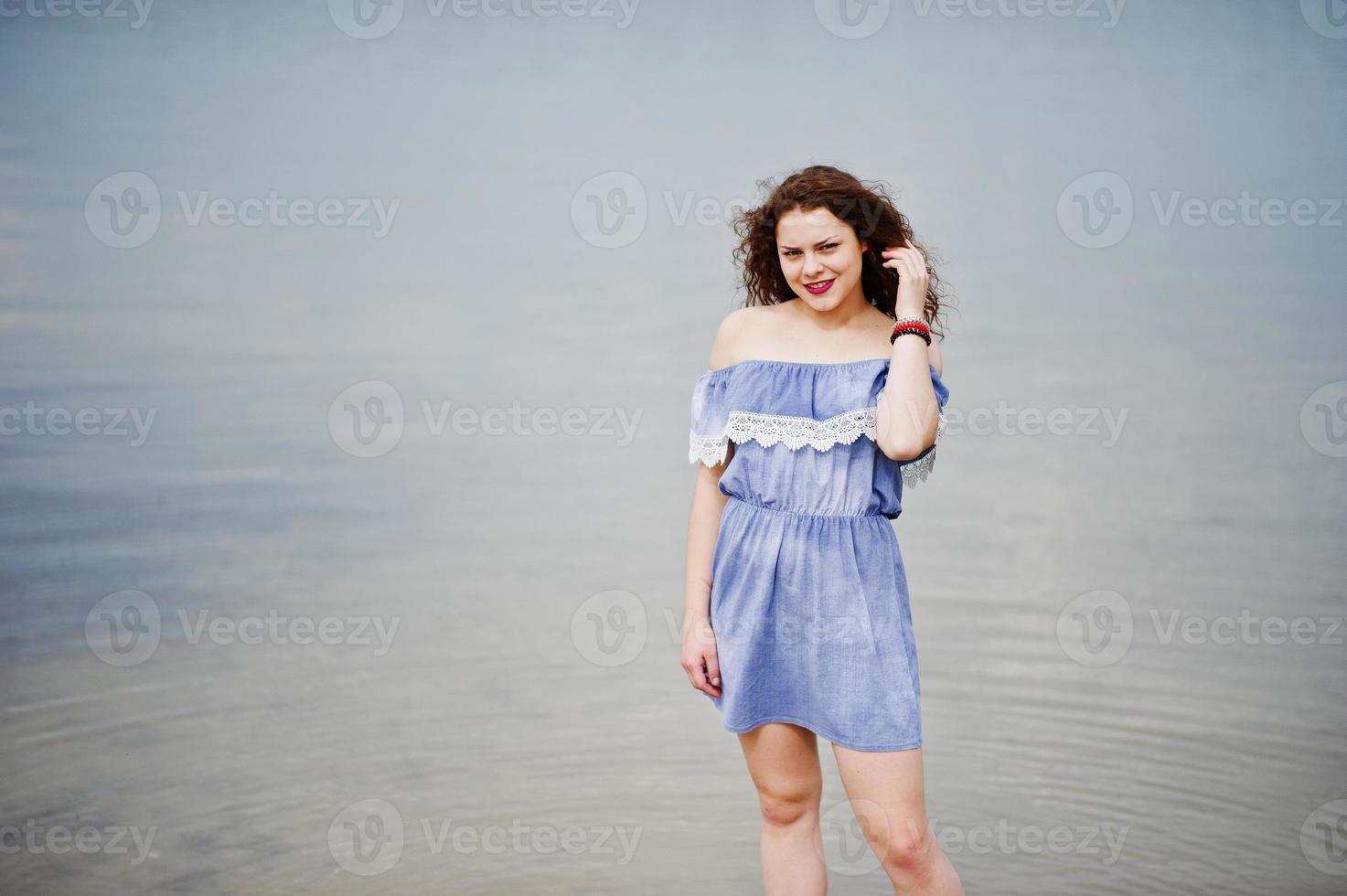Portrait of a beautiful girl with curly hair wearing marine blye dress stands in the water. photo