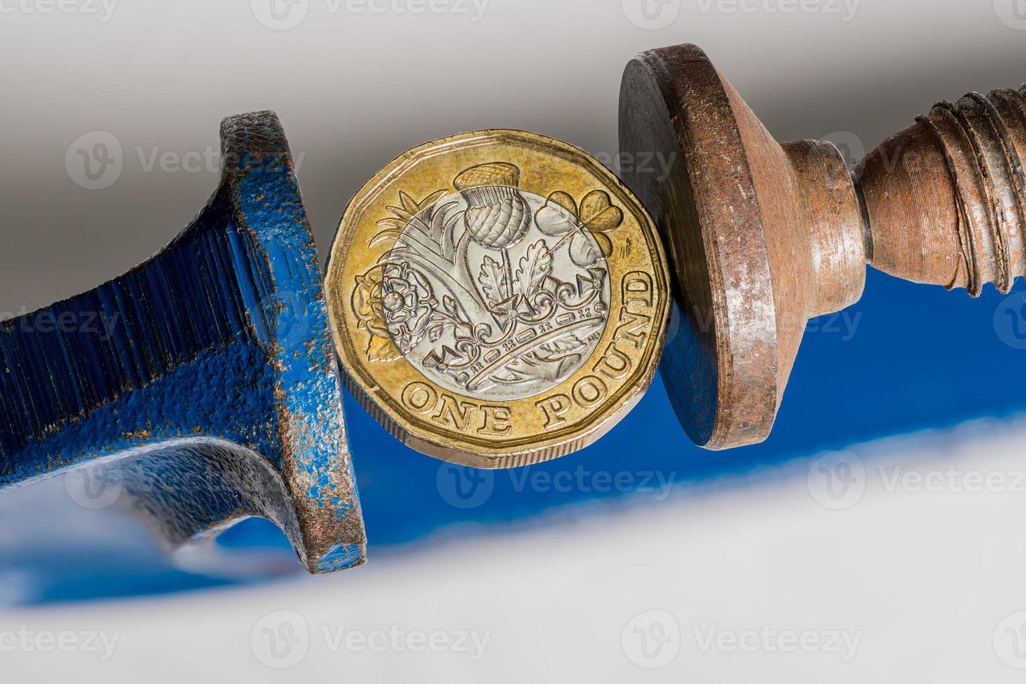 UK one pound sterling coin being squeezed between the solid jaws of a vise or vice photo