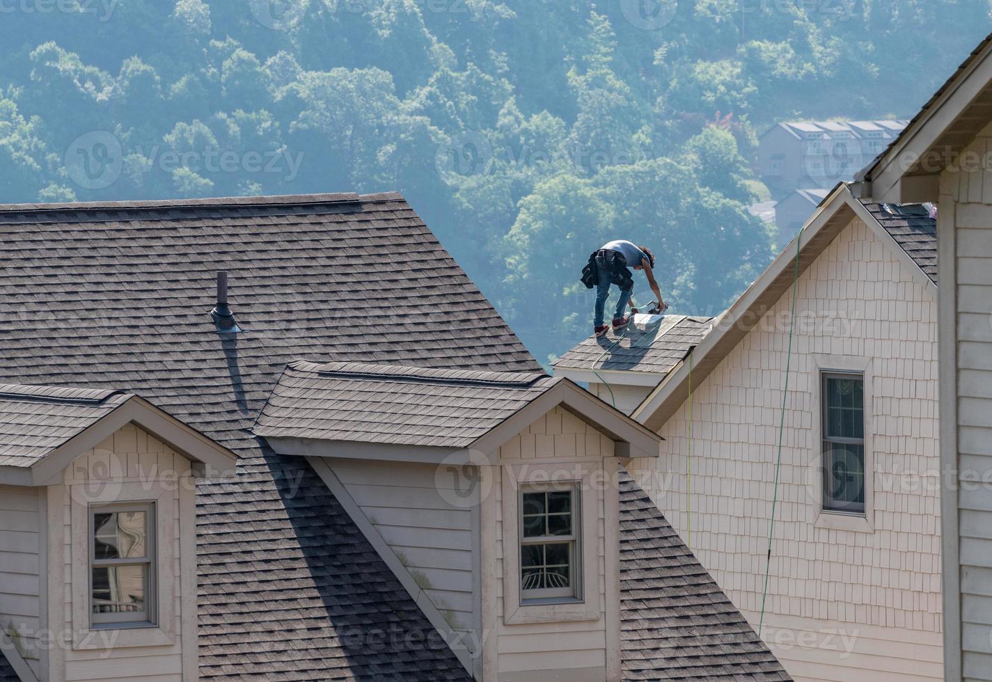 Young roofing contractor nailing shingles on a roof high above the ground photo