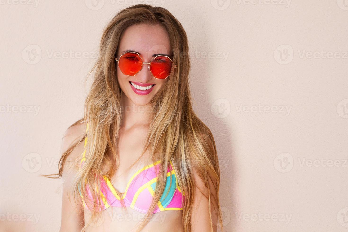 cerca de una chica feliz con gafas de sol rosas aisladas. vacaciones de verano y fin de semana divertido. concepto de verano. mujer joven sonriente en traje de baño de moda. enfoque selectivo. traje de playa de verano. copie el espacio foto