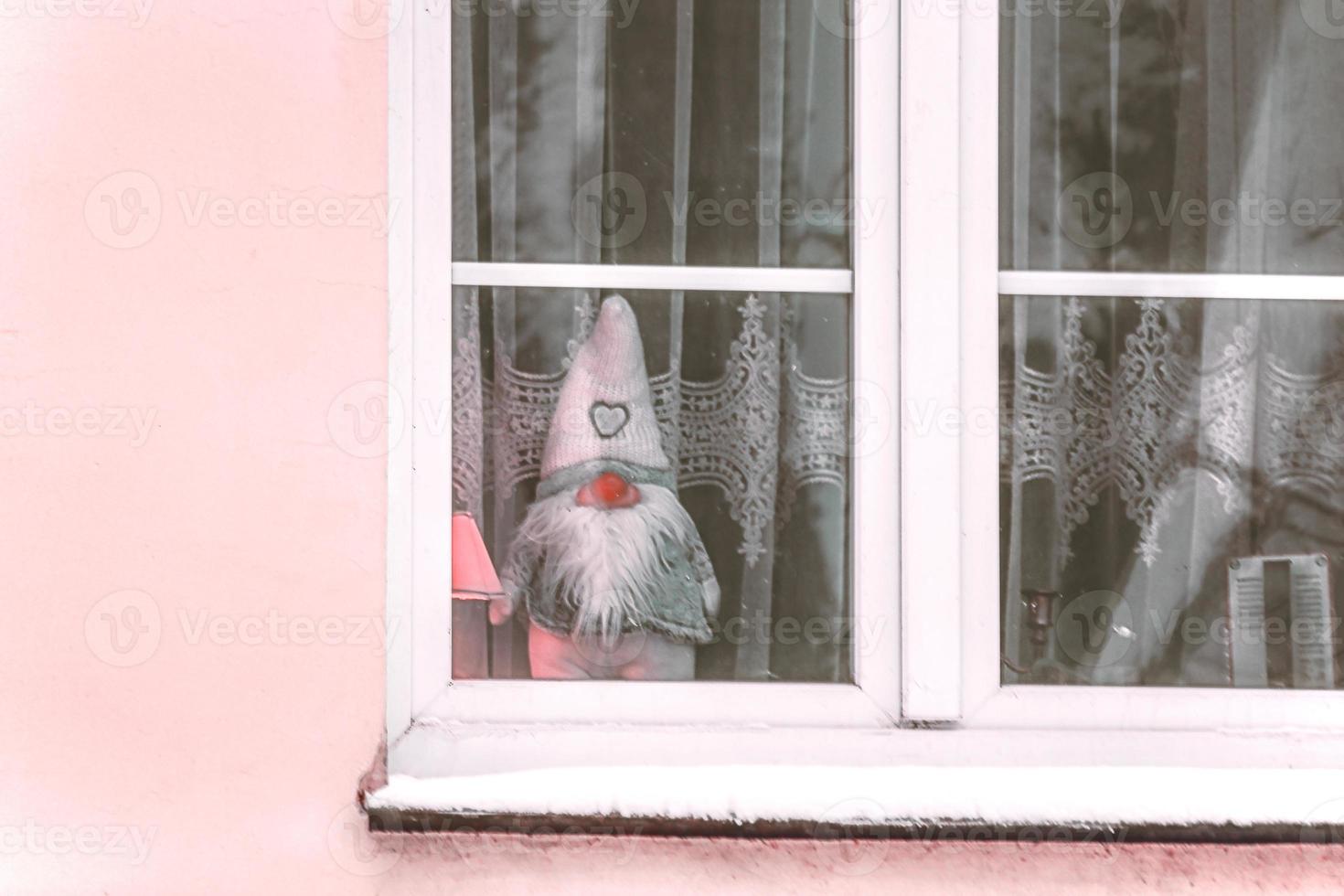 gnomo de juguete de fieltro parado detrás de la ventana con cortina de tul blanco en un edificio de pared rosa foto